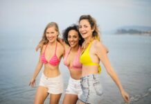 Travel-Trends-Studie – Der klassische zweiwöchige Sommerurlaub stirbt aus Drei junge Frauen in Shorts und Bikinioberteilen am Strand. Sie lachen
