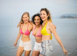 Travel-Trends-Studie – Der klassische zweiwöchige Sommerurlaub stirbt aus Drei junge Frauen in Shorts und Bikinioberteilen am Strand. Sie lachen