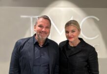 NOBILIS GROUP übernimmt Mehrheit an TMC Nordic Ein Mann und eine Frau, beide dunkel gekleidet, positionieren sich vor einer grauen Wand.