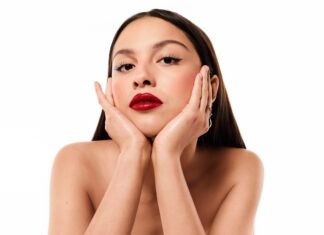 Olivia Rodrigo ist neue globale Lancôme-Markenbotschafterin Junge Frau mit rot geschminkten Lippen und langen dunklen Haaren, hat ihr Gesicht in den Händen aufgestützt und blickt in die Kamera.