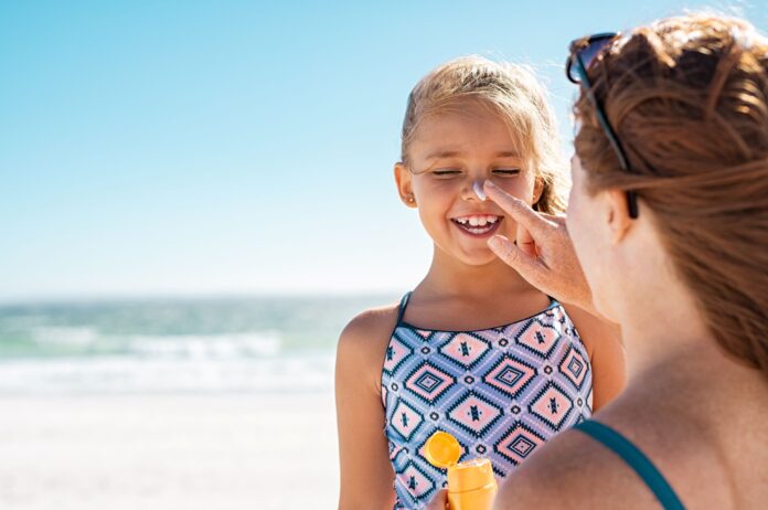 Mutter cremt das Gesicht ihrer mit Sonnenschutzmittel ein. die beiden sind am Strand am Meer.