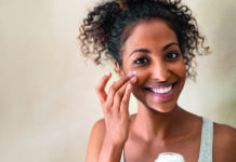 Sauber. Schön. Systemrelevant Smiling african girl with applying facial moisturizer while holding jar and looking at camera. Portrait of young black woman applying cream on her face isolated on beige background with copy space.