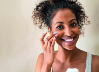 Sauber. Schön. Systemrelevant Smiling african girl with applying facial moisturizer while holding jar and looking at camera. Portrait of young black woman applying cream on her face isolated on beige background with copy space.