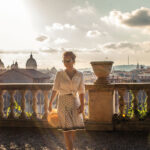 Salve Roma – Ein Besuch in der Ewigen Stadt Junge blonde Frau, sommerlich gekleidet auf einem antiken Balkon mit Aussicht über die Dächer von Rom