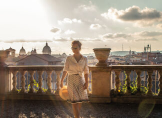 Salve Roma – Ein Besuch in der Ewigen Stadt Junge blonde Frau, sommerlich gekleidet auf einem antiken Balkon mit Aussicht über die Dächer von Rom