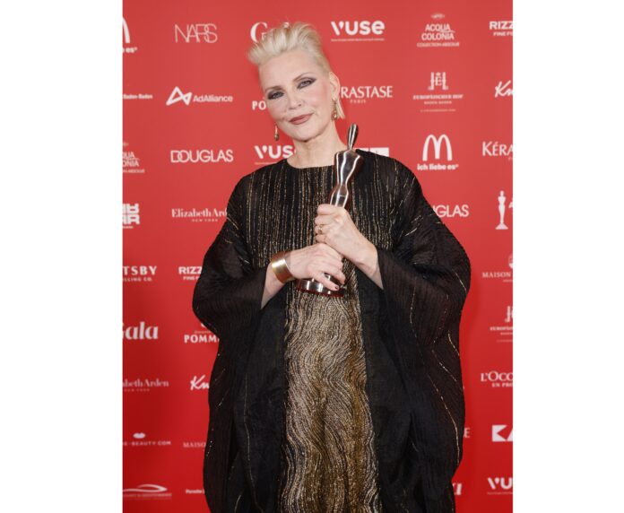 Frau mit kurzen blonden Haaren hält eine goldene Trophäe in der Hand. Sie steht vor einem roten Hintergrund