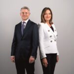 Die Müller Unternehmensgruppe richtet ihre Führungsstruktur neu aus Ein Mann im dunklen Anzug und eine Frau mit dunkler Hose und weißer Jacke stehen schräg nebeneinander, die Schultern berühren sich. Sie schauen in die Kamera.