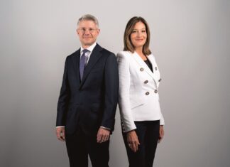 Die Müller Unternehmensgruppe richtet ihre Führungsstruktur neu aus Ein Mann im dunklen Anzug und eine Frau mit dunkler Hose und weißer Jacke stehen schräg nebeneinander, die Schultern berühren sich. Sie schauen in die Kamera.
