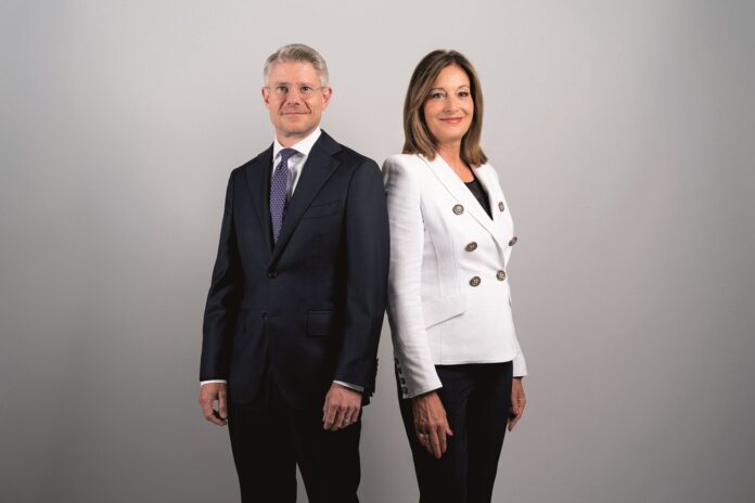 Ein Mann im dunklen Anzug und eine Frau mit dunkler Hose und weißer Jacke stehen schräg nebeneinander, die Schultern berühren sich. Sie schauen in die Kamera.