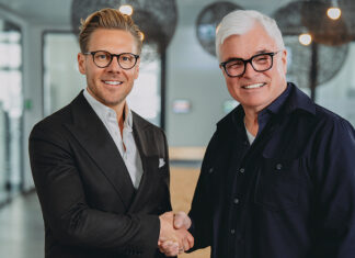 Strategische Allianz Zwei Männer mit Brillen schütteln sich die Hände und schauen in die Kamera.